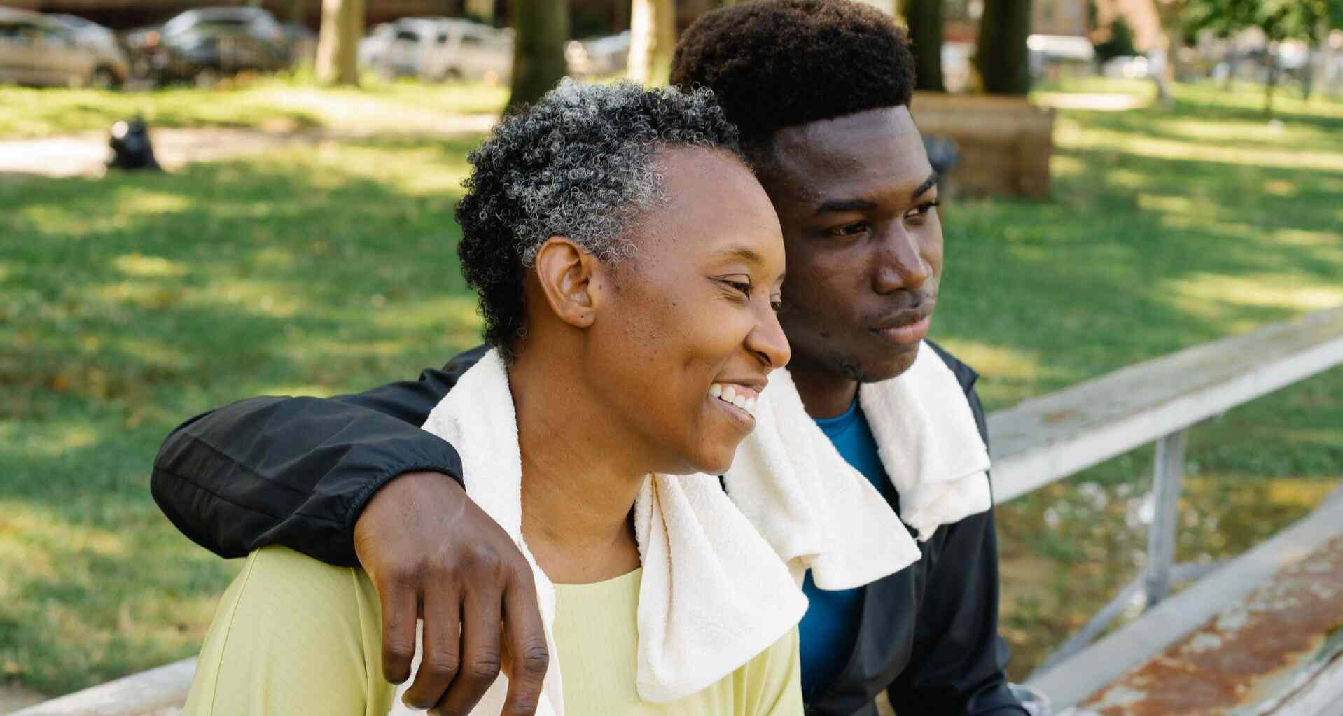 Mother and son sit together on a park bench after a workout, son's arm is around her shoulder