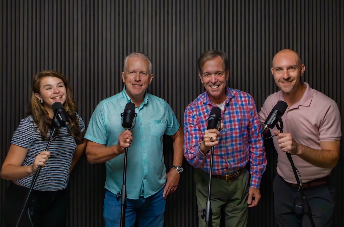 Four people stand holding microphones in front of a dark slatted studio wall