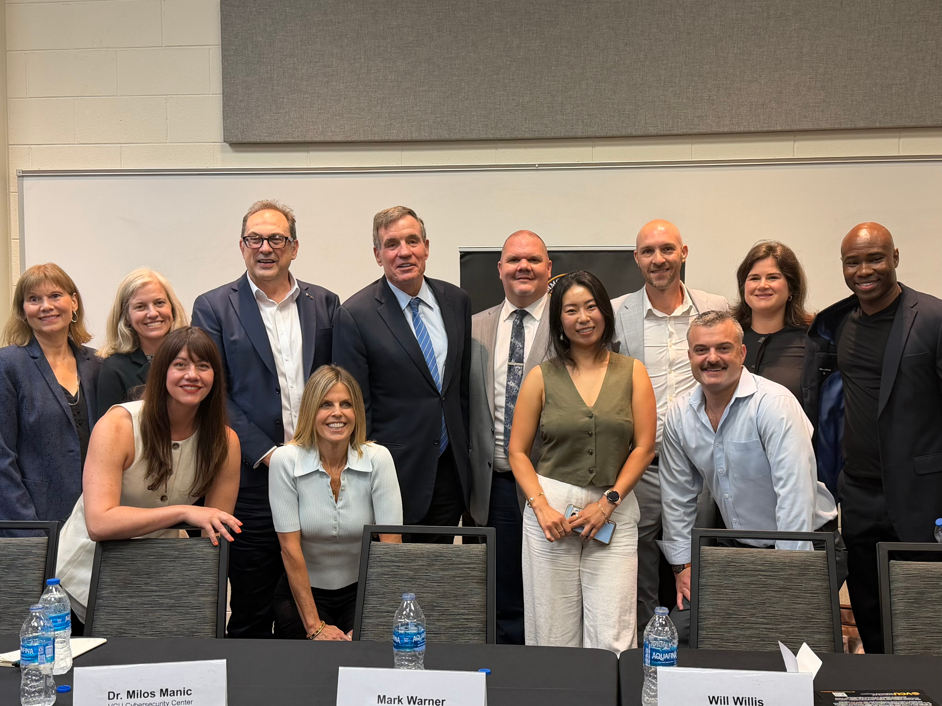 Sen. Mark Warner stands with twelve AI Ready RVA roundtable participants behind a table with name cards at VCU.